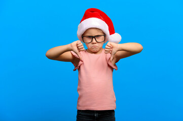 Obraz premium Happy new year concept. Upset caucasian girl wearing glasses and santa claus red hat shows thumb down on blue isolated background