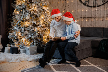 Father and son kid children are sitting near the Christmas tree and reading a book