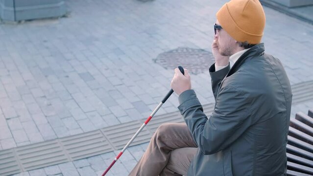 Young blind man with smartphone sitting on bench in park in city, calling