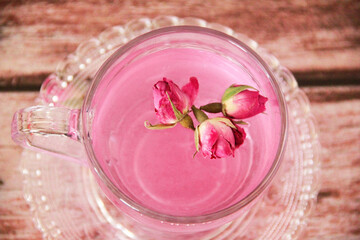 Tea made from pink rose petals in a transparent glass mug and small flower buds on a wooden background