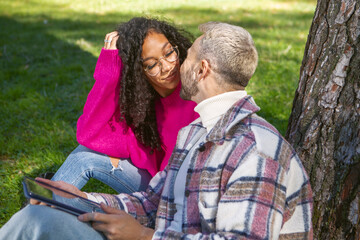 Fototapeta premium Smiling and happy couple sitting on the grass in the park looking into each other's eyes. Using technology
