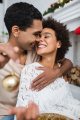 cheerful african american man embracing sexy girlfriend while holding blurred christmas ball