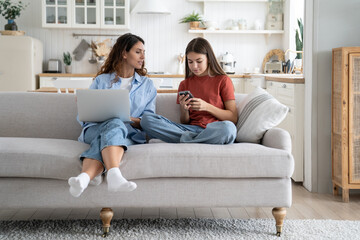 Friendly optimistic mother and daughter sits on sofa use modern electronic gadgets. Good-natured...