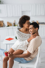 sensual african american woman hugging boyfriend sitting in kitchen with red cup