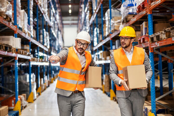 Two warehouse men in suits hold boxes with goods and relocating it while one of them is pointing where to put them.