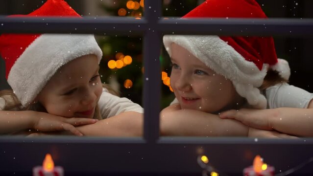 Close up of little girls in red santa hat on background of shining Christmas tree fun look at snow out window in evening. Happy children outside winter window