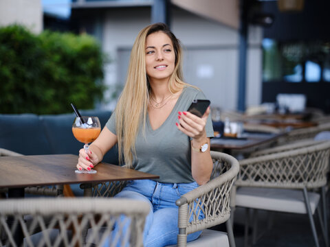 A happy woman sits in a cafeteria, texing messages and enjoying juice while smiling at the phone.