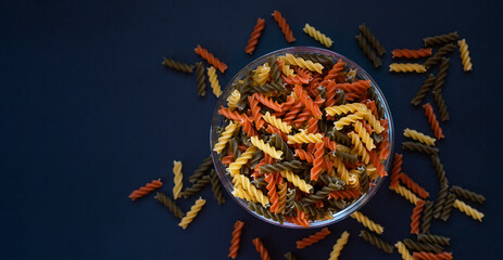 Tricolor organic fusilli paste in glass bowl on dark background. Top view. Close up. Copy space....