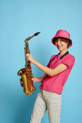 Fototapeta na wymiar Portrait of cheerful teen girl with curly brown short hair posing in pink panama and playing saxophone isolated over blue background. Musician