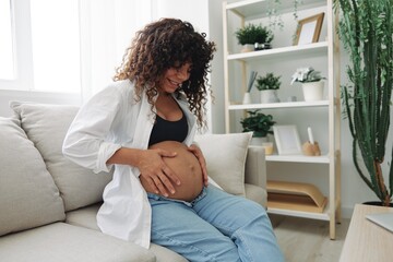 Pregnant woman smile and happiness sits on the couch freedom and strokes her belly feels kicks with...