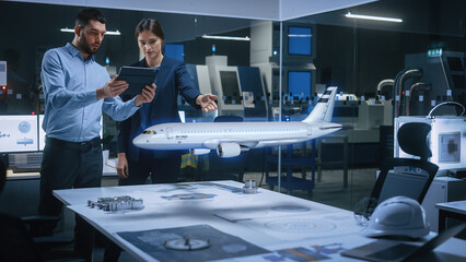 Aeronautics Factory Office Meeting Room: Chief Engineer Holds Tablet Computer, Showing Augmented Reality Airplane to a Female Project Manager, They Test Aerodynamics. Modern Industry 4.0 Research. - obrazy, fototapety, plakaty