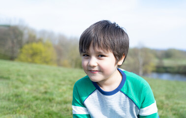 Portrait Young boy looking at camera with smiling face, School kid sitting  alone on green grass in garden, Positve Cute Child playing outdoor with blurry green nature in the park on SPring or Summer