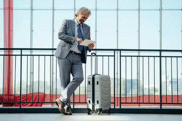 Happy business man in elegant gray suit with wireless earphones having online meeting on his digital tablet while standing in terminal hall with his suitcase