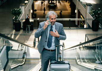 Fototapeta na wymiar Smiling Mature Businessman Talking on a Mobile Phone while Moving Up Escalator with his Luggage at the Airport