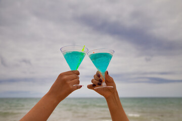 Detail of two hands raising glasses in the air. They are toasting for the good things that happen. The photo is taken from below and you can see the sky and the sea in the background.