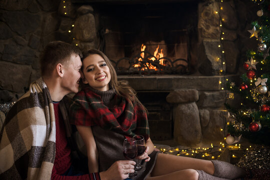 Young heterosexual couple in love in sweaters and plaids near the fireplace. A guy and a girl celebrate Christmas by the fireplace with New Year's garlands and a Christmas tree in a warm cozy atmosphe