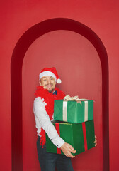 Smiling man wearing Santa hat holding Christmas gift boxes on the red arch background. New Year style