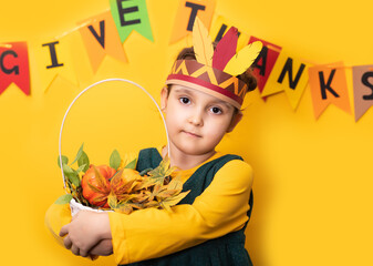 Thanksgiving day concept. Cute little kid girl in paper native american hat holds basket with harvest gifts on yellow background. - obrazy, fototapety, plakaty