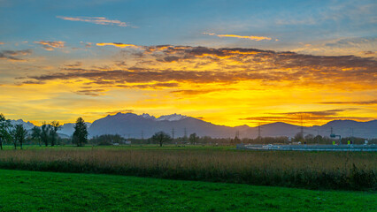 Sonnenuntergang im Rheintal, mit Wiesen und Felder, Bäumen und Schweizer Bergen im Hintergrund. Föhn mit Wolken und blau, gelb, orange und rotem Himmel 