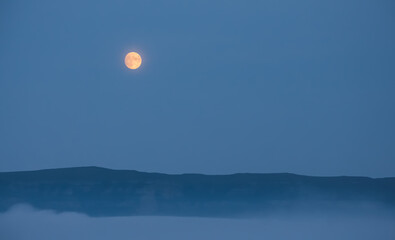 Full moon in the mountains in the early morning with a mountain cliff and dense clouds in the valley, orange moon in the autumn morning in the mountains