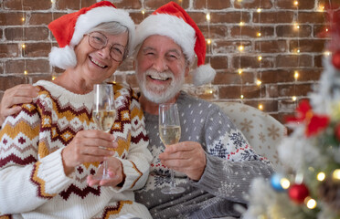 Cheerful senior adult couple at home enjoying christmas together in december wearing santa hats....