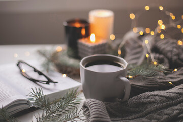 Winter home cozy concept. Mug with coffee, open book, warm sweater, candles and fir tree. Wellbeing, relaxing concept
