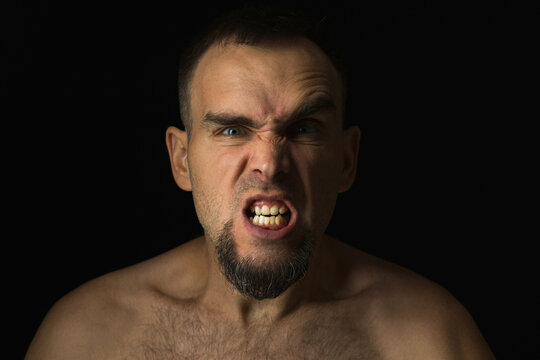 Portrait of an angry man with open mouth and frowning nose and raised eyebrow. Caucasian man with shirtless beard on black background. Anger, aggression and rage on a human face.