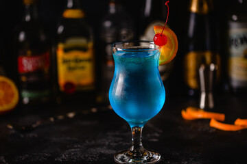 Glass of blue lagoon cocktail decorated with orange and cherry at festive bar counter background.