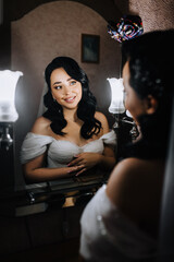 A beautiful, smiling, curly-haired bride model in a white dress stands in front of a mirror, reflecting, illuminating with a lamp. Wedding photography, portrait.