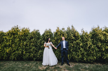 A stylish groom in a blue suit and a beautiful bride in a white long dress are standing in a park, garden near green thujas, cypress. Wedding photography, portrait.