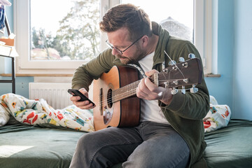 Man with an acoustic guitar is learning a new melody while looking at notes on a smartphone.