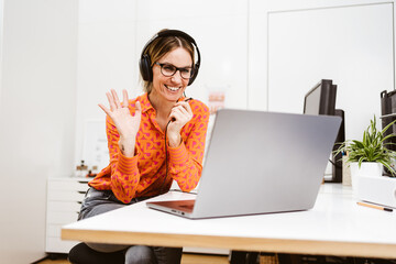 Young laughing business woman with headphones and laptop says hello in online meeting