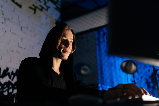 Close-up view of dangerous hacker man in sweatshirt hacking online web site or engaging hacking into security systems, using laptop and desktop computer in dark basement room with blue neon lights.