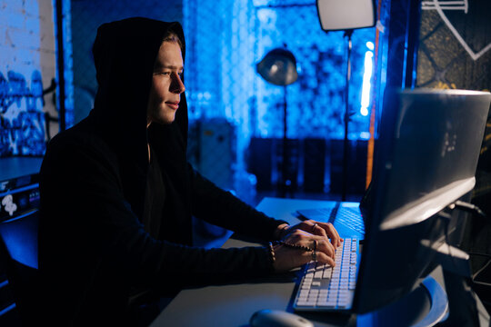 Side view of hidden hacker man wearing sweatshirt with hood engaged in hacking into security systems, sitting in dark room with blue neon lights. Concept of cyberattack, malware and cyber security.