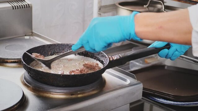 Chef in a professional restaurant kitchen stirs and turns meat on hot pan surface. Professional cook in blue gloves frying chicken liver in iron skillet. Cooking, preparing ingredients for customer