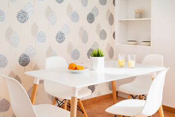 White kitchen table with two glasses of orange juice, a split fresh orange, green natural plant and leaf wallpaper background, in grey, blue and white
