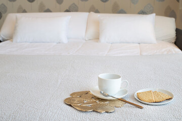Bedroom with white bed. Small white coffee cup, coffee plate with cookies and gold trivet with seashell decoration on gray blanket