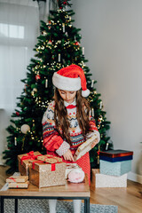 A little girl in Santa hat and mom's festive knitted sweater packing gift boxes on the Christmas tree background. Christmas mood concept. Preparing or opening presents. Vertical card. Selective focus.