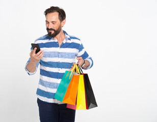 Attractive beard man looking into holding smartphone and bunch of colored shopping bags isolated over white background studio, shopping, communication and finance concept.