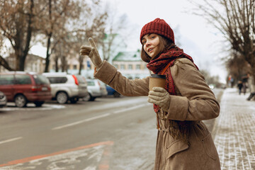 Woman wearing winter clothes with cup of coffee to go waving hand and catching taxi on street in city 