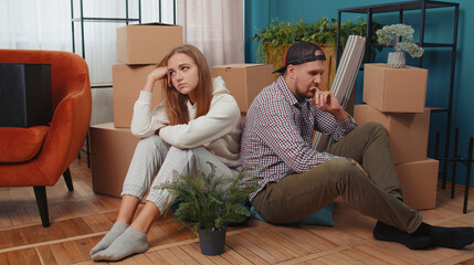 Sad tired married couple man woman leave house sit on floor near cardboard boxes with belongings...
