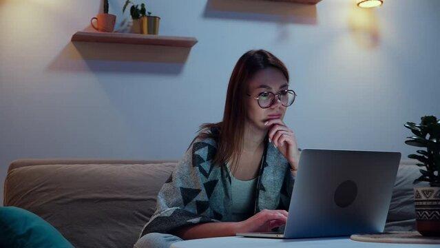 Young smiling woman using laptop at night, working overtime at home sitting on sofa, flexible working hours