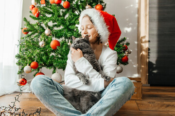 A girl in a santa claus hat hugs her gray cat on the eve of Christmas next to the Christmas tree....