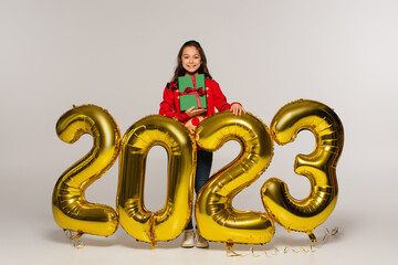 full length of happy kid in  sweater holding christmas present near balloons with 2023 numbers on...