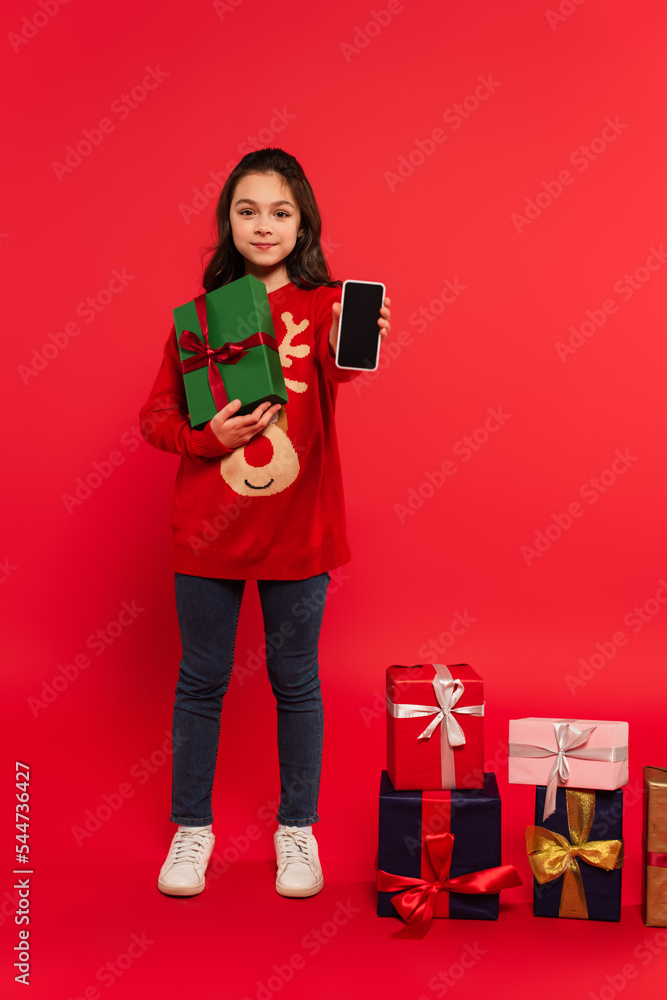 Wall mural full length of happy child in knitted sweater showing smartphone with blank screen near Christmas presents on red