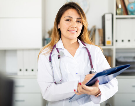 Positive latino woman doctor in white coat standing in office with clipboard making notes