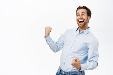 Handsome triumphing man fist pump, celebrating victory or achievement, smiling pleased, standing over white background - obrazy, fototapety, plakaty