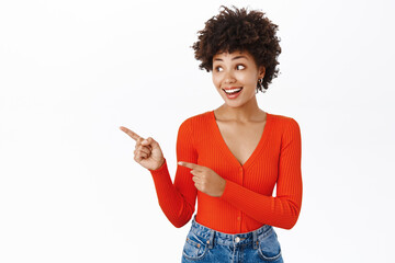 Smiling african american woman in stylish outfit, pointing finger left, showing promotion text on copy space, empty banner, stands over white background