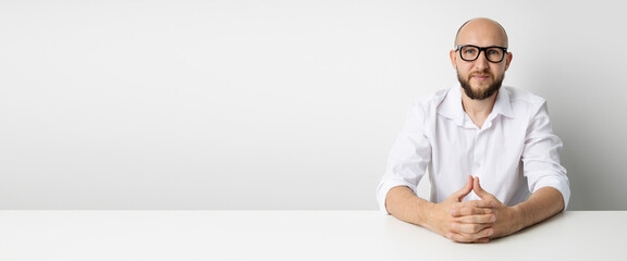 Young man holding hands folded while sitting at table on white background. Banner