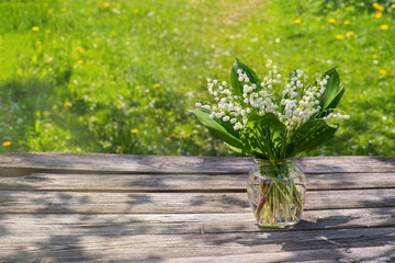 Beautiful bouquet of lilies of valley in glass transparent jar of water on old wooden table on green background. Bouquet of lilies of valley in vase on old table.